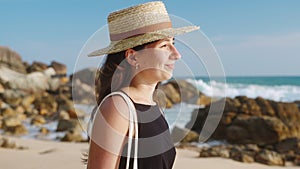 Woman in straw hat enjoys picturesque sea sunset. Solo traveler strolls along sandy shore. Waves crash against rocks