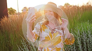 Woman in a straw hat and flowery dress saying hello having a video call in the field at sunset