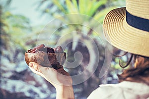 Woman in a straw hat holding in a hand royal dates fruit in a bowl of coconut in a palm tree grove