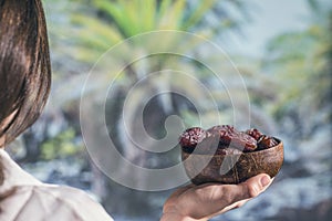 Woman in a straw hat holding in a hand royal dates fruit in a bowl of coconut in a palm tree grove