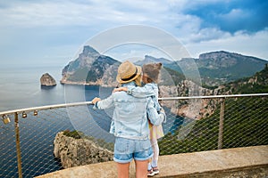 A woman in straw hat hugging her little daughter, while gazing at a breathtaking view from Mirador de El Colomer in Mallorca.