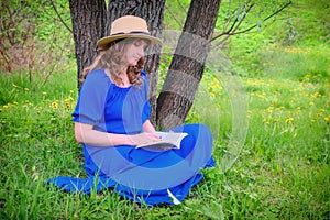 A woman in a straw hat reading a book on the grass sitting under a tree