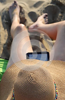 woman with straw hat while reading an e-book on the beach and re