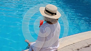 woman in a straw hat relaxes near the swimming pool, drinks a pink cocktail and dangles her legs in the water. Summer