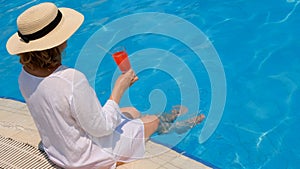 woman in a straw hat relaxes near the swimming pool, drinks a pink cocktail and dangles her legs in the water. Summer