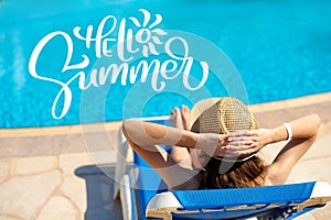 Woman in a straw hat relaxing on a deck-chair near a luxurious blue pool and letters Hello Summer, concept time to