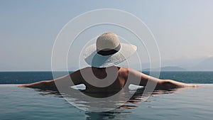 Woman with straw hat relaxing in infinity swimming pool with sea view at luxury resort. summer vacation