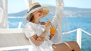 Woman with a straw hat settles in a gazebo on a wooden sea pier. Sipping her drink, she finds peace watching sea