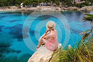 A woman in straw hat sitting on a cliff, looking towards Cala Gat beach in Mallorca