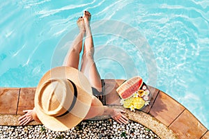 Woman in straw hat sitting on swimming pool side  with plate of tropical fruits- camera top view