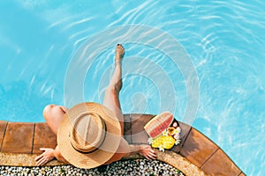 Woman in straw hat sitting on swimming pool side  with plate of tropical fruits. Top view shot