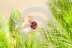 Woman in a straw hat sitting under coconut palm tree branches holding in a hand royal dates fruit in a bowl on the sand of beach.