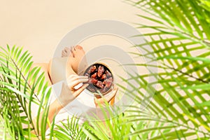 Woman in a straw hat sitting under coconut palm tree branches holding in a hand royal dates fruit in a bowl on the sand of beach.