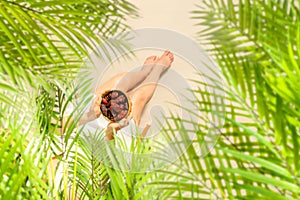 Woman in a straw hat sitting under coconut palm tree branches holding in a hand royal dates fruit in a bowl on the sand of beach.