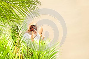 Woman in a straw hat sitting under coconut palm tree branches holding in a hand royal dates fruit in a bowl on the sand of beach.