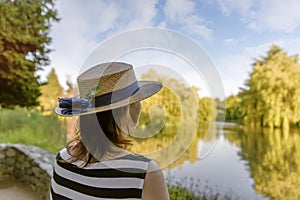 woman in a straw hat, standing in a park by the pond and admiring the nature