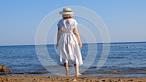 woman in a straw hat and white dress walks along the seashore and admires the blue sea on a sunny summer day, enjoying