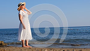 woman in a straw hat and white dress walks along the seashore and admires the blue sea on a sunny summer day, enjoying