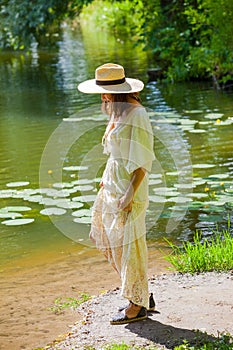 Woman in a straw hat and white summer dress on the shore of an old pond