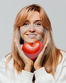 Woman with strawberry blonde hair hold red heart, smiling,looking at camera, closeup, isolated