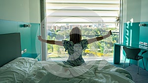 Woman stretching and sitting on the bed after waking up in front of a large window with a view.