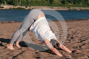Woman stretching yoga on the beach by the river in the city. Beautiful view. Adho mukha svanasana pose.