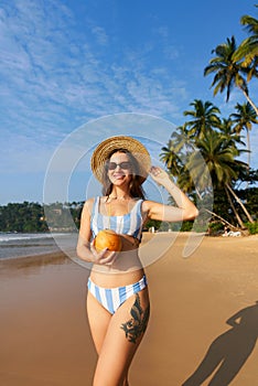 Woman in striped bikini holds fresh coconut on sunny beach. Solo traveler enjoys tropical seaside, healthy drink. Palm