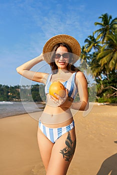 Woman in striped bikini holds fresh coconut on sunny tropical beach. Fit female in hat enjoys summer, drinks healthy