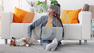 Woman stroking dog and working with laptop, sitting by sofa in apartment interior spbi.