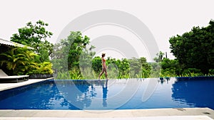 Woman strolls by luxe villa infinity pool, enjoys lush tropical landscape. Side view of female in swimsuit walking