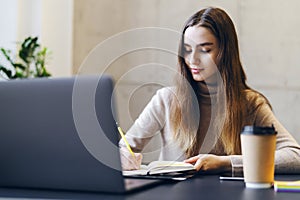 Woman student sitting in front of laptop, streaming webinar online, making notes in copybook. Young specialist doing research on