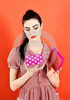 Woman in stylish dress opens present. Girl with unsatisfied face, make up and pink pack. Lady with gift isolated on red