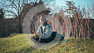 A woman in a stylish hat uses her phone while sitting on the grass in a park