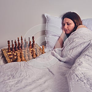 A woman is suffering from insomnia on a bed next to a chess board. A lonely adult woman lies in bed with a sad face
