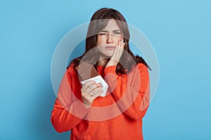 Woman suffering from toothache after eating chocolate  frowning her face  covering cheek with palm  female with dark hair wearing