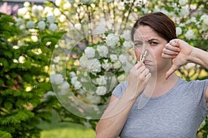 A woman suffers from a clothespin on her nose and shows a thumbs down on a walk in a flowering park. Allergy.