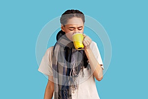 Woman suffers from cough and bad cold, standing wrapped in scarf, drinks hot beverage, cures disease