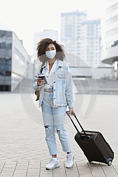 Woman with suitcase holding passport and tickets, girl wearing protective face mask in a city. Business travel during pandemic
