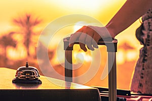 Woman with suitcase ringing hotel service bell with sea and palm tree view on sunset. Travel concept. 24-hour beach hotel front