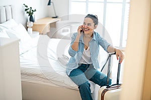 Woman with a suitcase sitting on bed in hotel room and using phone