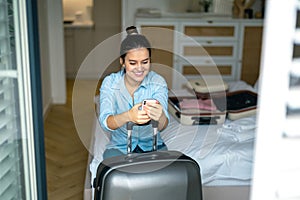 Woman with a suitcase sitting on bed in hotel room and using phone