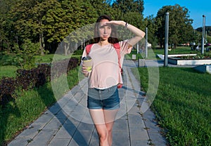 woman in summer in a city park looks into distance, covers herself with her hand from sun, a meeting and a date, a cup