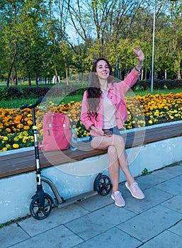 Woman in summer in a city park waves her hand, happy smiles rejoices, meeting and a date, scooter shop bag and bottle