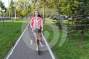 woman in summer in city rides a scooter on a bicycle path, in motion, background road trees green grass and spruce, pink