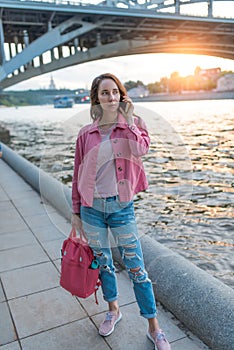 A woman in summer in city stands by river bank, making a phone call, dating and meeting on street, traveling and