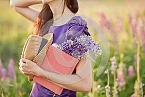 Woman in summer field with bouquet of wildflowers - lupines, hat and pink notebook. Closeness to nature, self-discovery