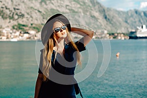 A woman in sun glasses and a hat stands against the backdrop of the sea and mountains. Vacation and outdoor recreation. Girl with