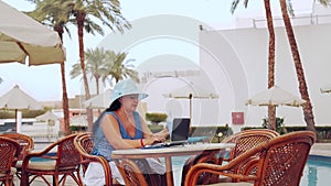 A woman in a sun hat sits at a table by the pool and works with a laptop.