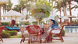 A woman in a sun hat sits at a table by the pool and works with a laptop and drinks a cocktail