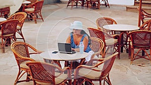 A woman in a sun hat sits at a table by the pool and works with a laptop and drinks a cocktail